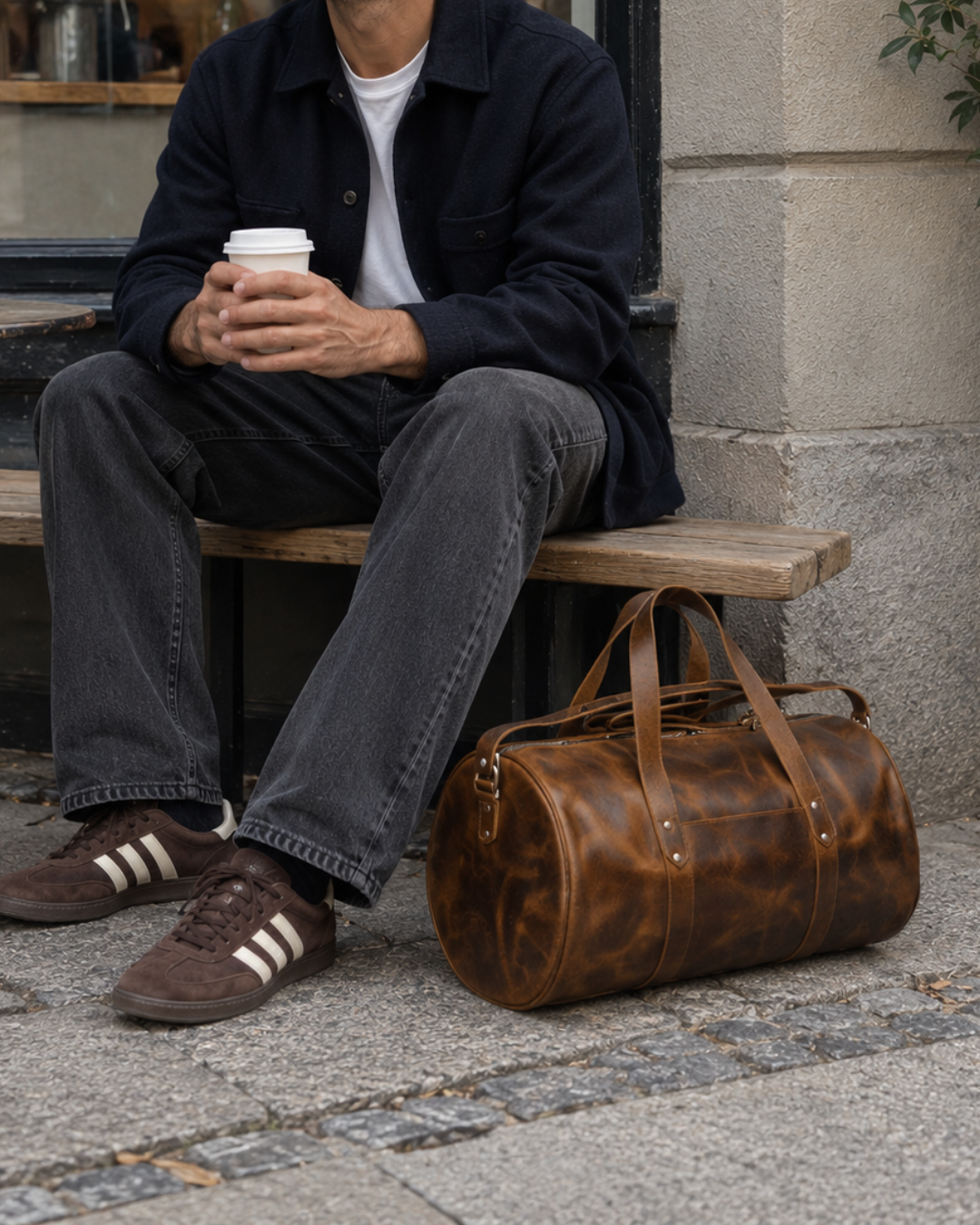 Laylowe Wells light brown leather weekender beside a man sitting outside a cafe with a takeaway coffee.