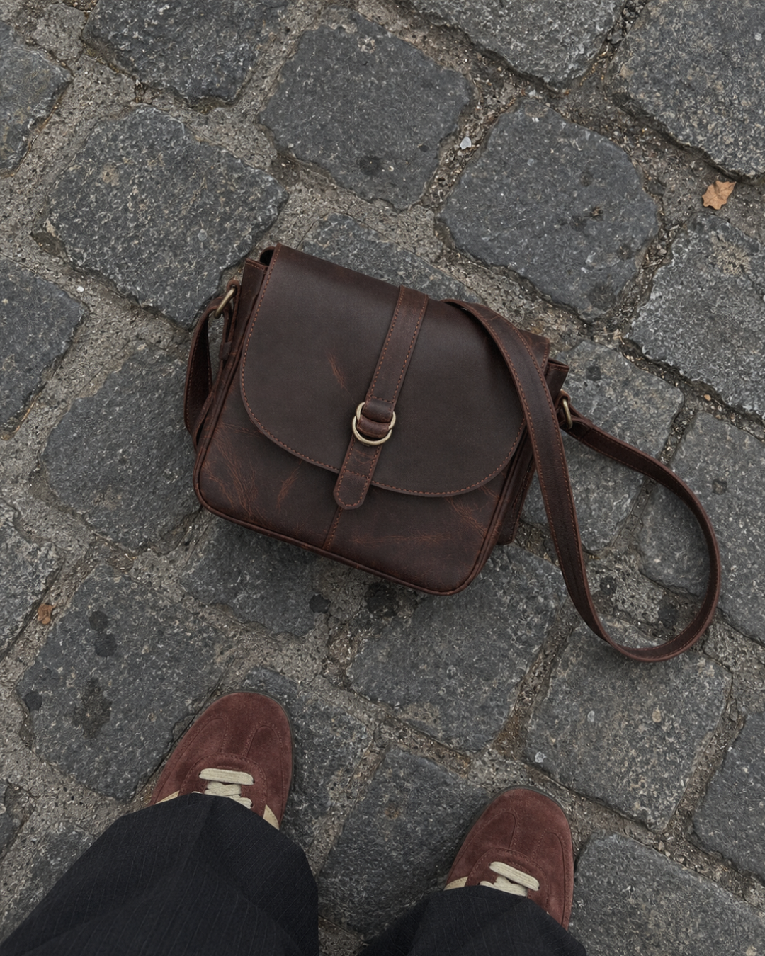 Top-down view of the Laylowe Cora dark brown leather satchel on cobblestones beside suede sneakers.
