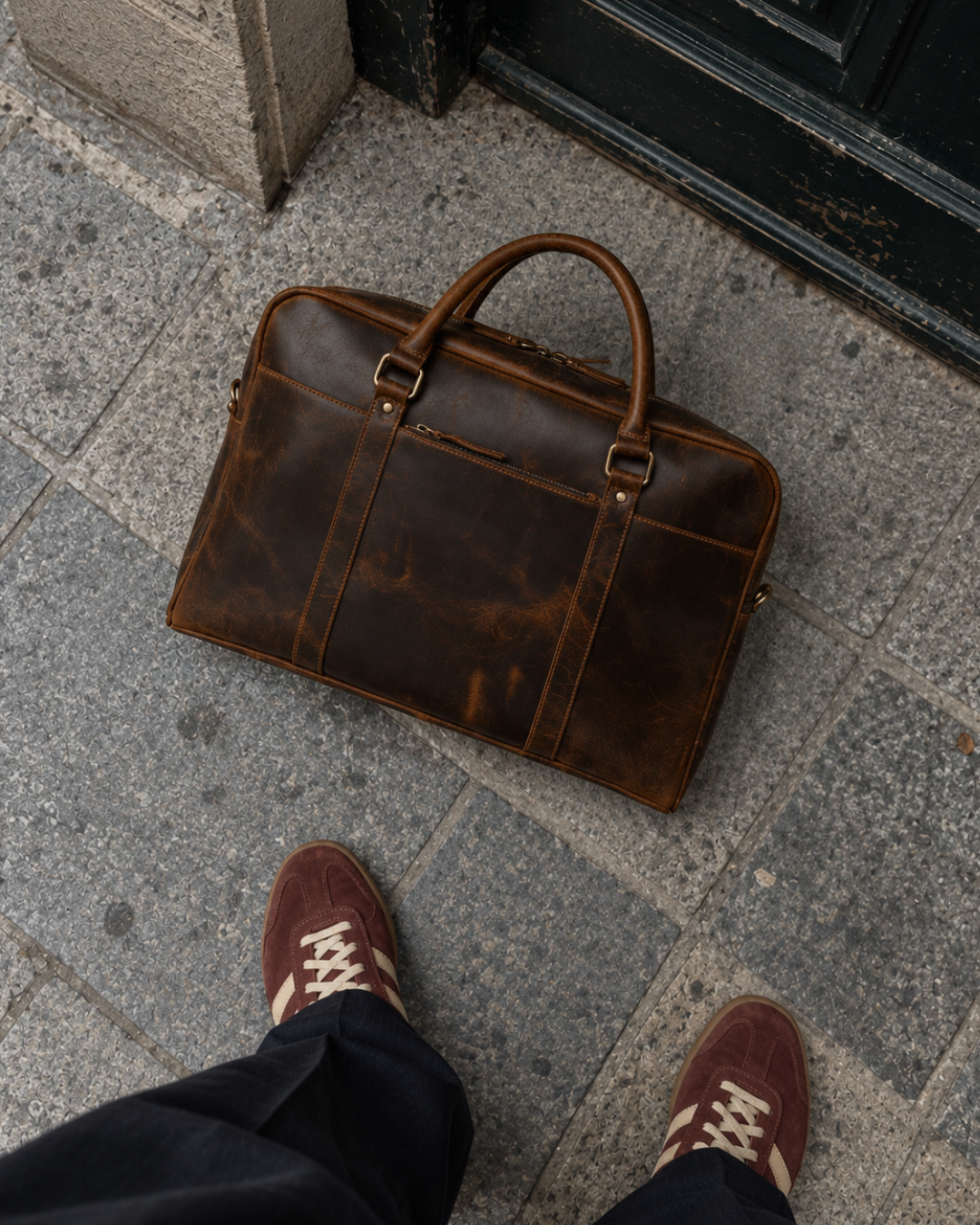 Top-down view of a Laylowe Reid brown leather briefcase on stone pavement beside brown suede sneakers.