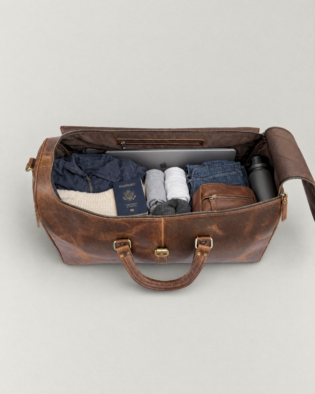 Top view of an open Laylowe Rhodes light brown leather duffle packed with a laptop, clothes, passport, pouch and bottle.