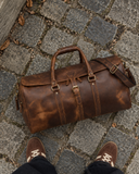 Top-down view of a Laylowe Rhodes light brown leather travel bag on cobblestones beside suede sneakers.