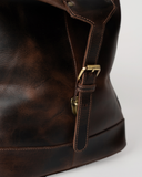Side detail of the Laylowe Hayes dark brown leather weekender, showing its buckle strap and curved leather panel.