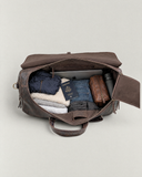 Open Laylowe Rhodes dark brown leather duffle packed with a laptop, passport, folded clothes, toiletry pouch and dark bottle.