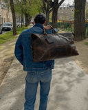 Man in a denim jacket carrying the Laylowe Hayes dark brown leather weekender over his shoulder on a tree-lined path.