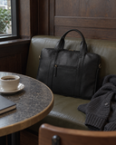 Laylowe Nolan black leather laptop case resting on a cafe bench beside a cup of coffee and folded knitwear.
