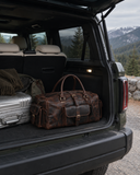 Laylowe Bennett dark brown leather travel bag beside silver luggage in an open SUV trunk overlooking mountains.