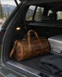Laylowe Wells light brown leather weekender in an SUV trunk beside a silver suitcase and knitwear, with mountains beyond.