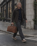 Man crossing a city street carrying a Laylowe Parker tan leather duffle bag by its rolled handles.