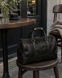 Laylowe Beck dark brown small leather duffle on a wooden cafe chair beside a table with coffee and flowers.
