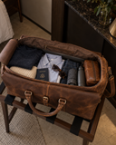 Open Laylowe Rhodes light brown leather travel bag on a luggage rack, packed with clothes, sunglasses, passport and bottle.