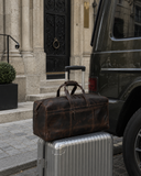 Laylowe Rowan dark brown leather duffle resting on a silver rolling suitcase beside an olive SUV.