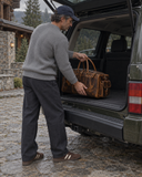Man loading a Laylowe Alden brown leather weekender into the open rear of an SUV outside a stone lodge.