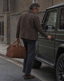 Man carrying a Laylowe Parker tan leather weekender while opening an olive SUV door on a city street.