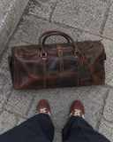 Overhead view of the Laylowe Holden dark brown leather travel bag on stone pavement beside brown sneakers.