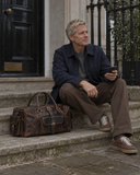 Man seated on stone steps beside a Laylowe Bennett dark brown leather travel bag with a front buckle pocket.