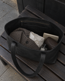 Open Laylowe Arden black leather tote on a wooden bench, holding folded clothes, a book and small accessories.