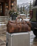 Laylowe Rhodes light brown leather travel bag on a silver suitcase beside an olive SUV outside a snowy mountain lodge.