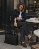 Woman seated outside a cafe with a Laylowe Arden black leather tote bag beside her chair.