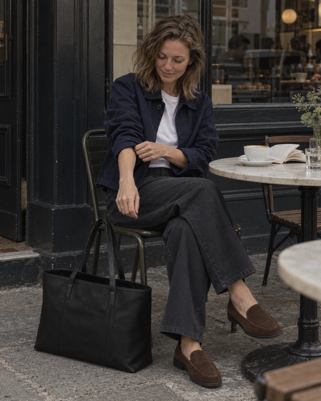 Woman seated outside a cafe with a Laylowe Arden black leather tote bag beside her chair.