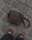 Top-down view of the Laylowe Cora dark brown leather satchel on cobblestones beside suede sneakers.