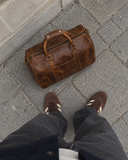 Overhead view of a Laylowe Beck light brown small leather duffle on stone pavement beside suede sneakers.