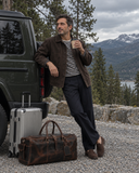 Laylowe Holden dark brown leather travel bag beside silver luggage and a man leaning against an SUV in the mountains.