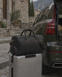 Laylowe Blake black leather duffle resting on a silver suitcase beside a green SUV on a stone street.