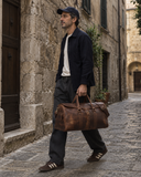 Man carrying a Laylowe Rhodes large light brown leather duffle along a narrow cobblestone street.