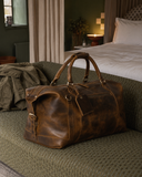 Laylowe Hayes light brown leather weekender resting on an olive knitted blanket at the foot of a bed.