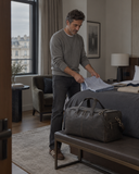 Man folding a shirt beside the Laylowe Ellis charcoal leather garment duffle on a bench at the foot of a bed.