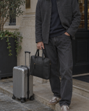 Man holding the Laylowe Nolan black leather laptop case beside a silver rolling suitcase on a stone pavement.