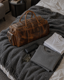 Laylowe Alden brown leather weekender on a bed beside folded clothes, a glasses case and fragrance bottle.