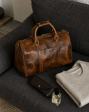 Laylowe Beck light brown small leather duffle on an armchair beside a folded sweater, glasses case and fragrance.