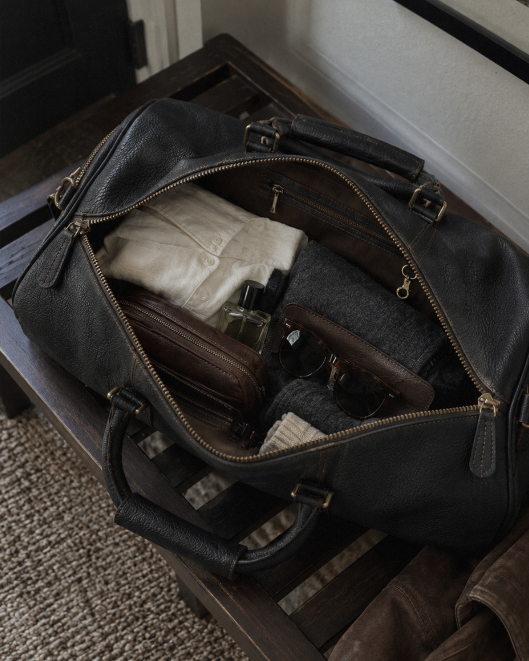 Laylowe Blake black leather duffle open on a luggage rack with folded knitwear, a toiletry pouch and sunglasses.