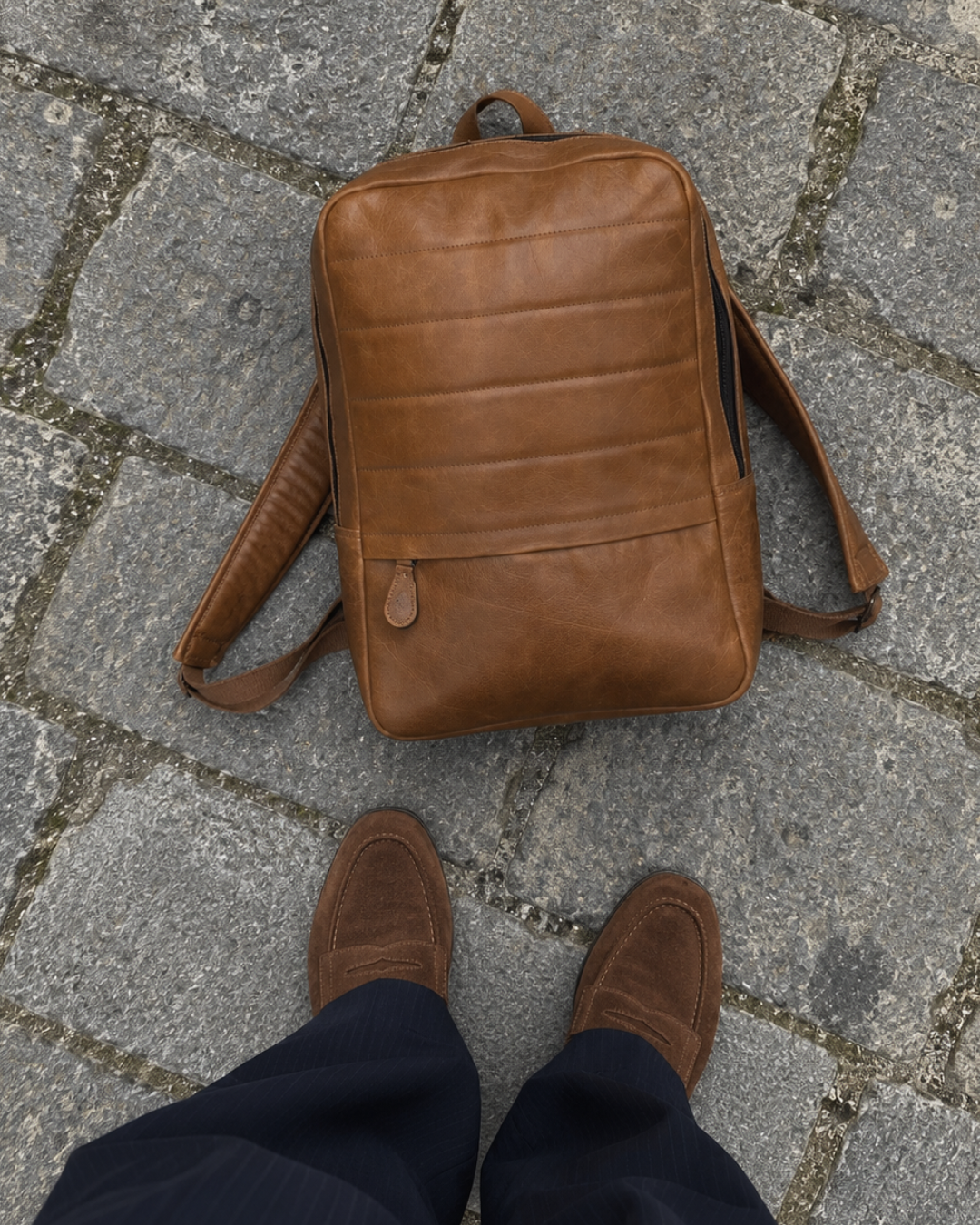 Overhead view of the Laylowe Lane tan leather backpack on stone pavement beside brown suede shoes.