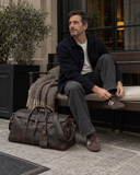 Laylowe Rhodes dark brown leather travel bag beside a man adjusting his shoe on an outdoor cafe bench.