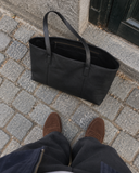 Top-down view of a Laylowe Arden black leather tote bag on cobblestones beside brown suede loafers.