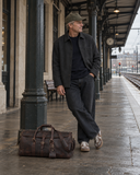 Laylowe Rhodes dark brown leather duffle on a train platform beside a man waiting beneath the station canopy.