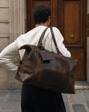 Laylowe Hayes dark brown leather weekender carried over one shoulder by a man standing at a wooden door.
