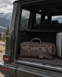Laylowe Rhodes dark brown leather travel bag and a silver suitcase in an open SUV trunk overlooking the mountains.