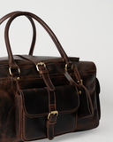 Close-up of the Laylowe Bennett dark brown leather travel bag with a buckled front flap pocket and rolled handles.