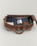 Top view of an open Laylowe Rhodes light brown leather duffle packed with a laptop, clothes, passport, pouch and bottle.